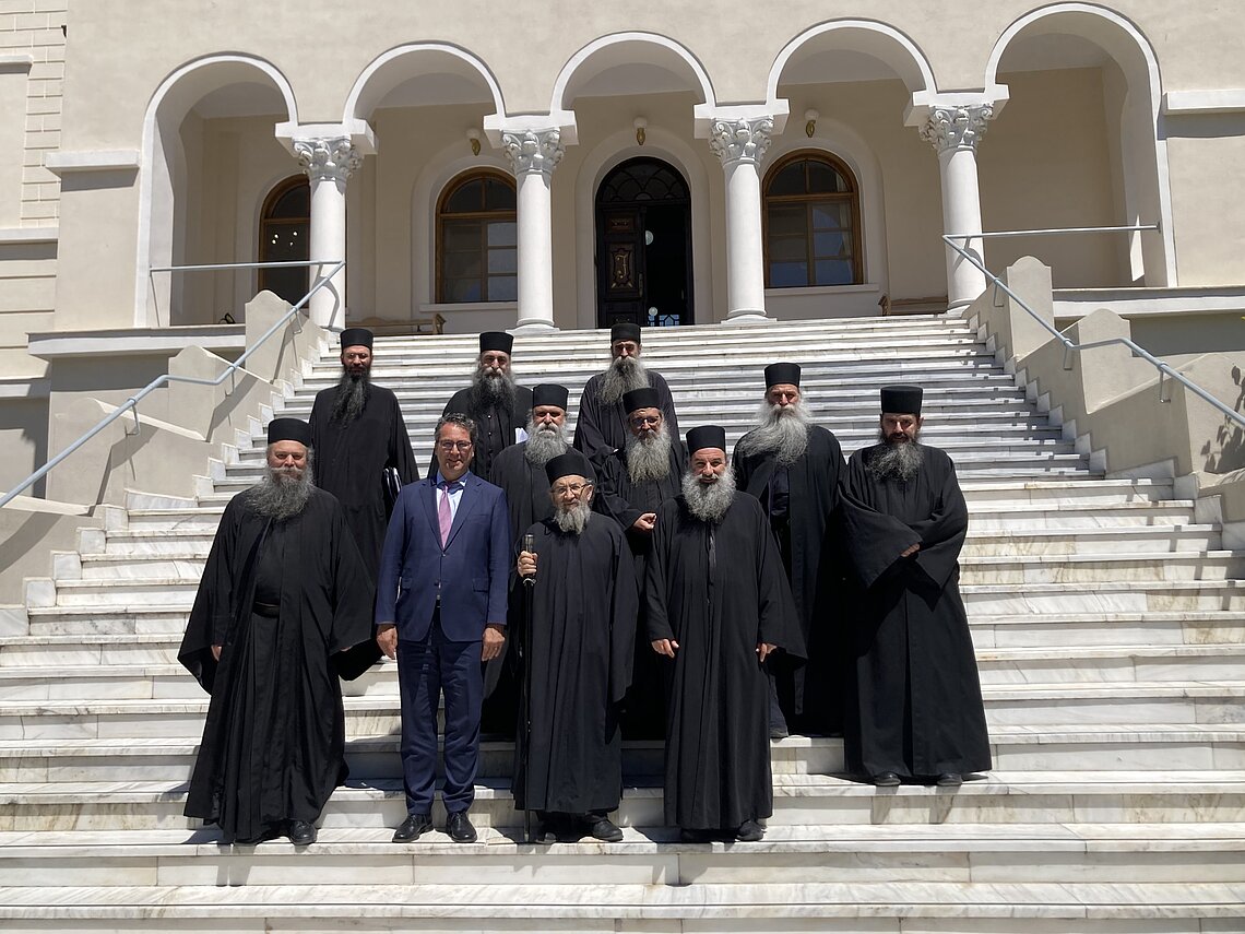 Eine Gruppe von Männern, die auf einer großen Treppe vor einem Gebäude mit Säulen und Bögen stehen. Die meisten tragen lange schwarze Gewänder, einer von ihnen trägt einen blauen Anzug.