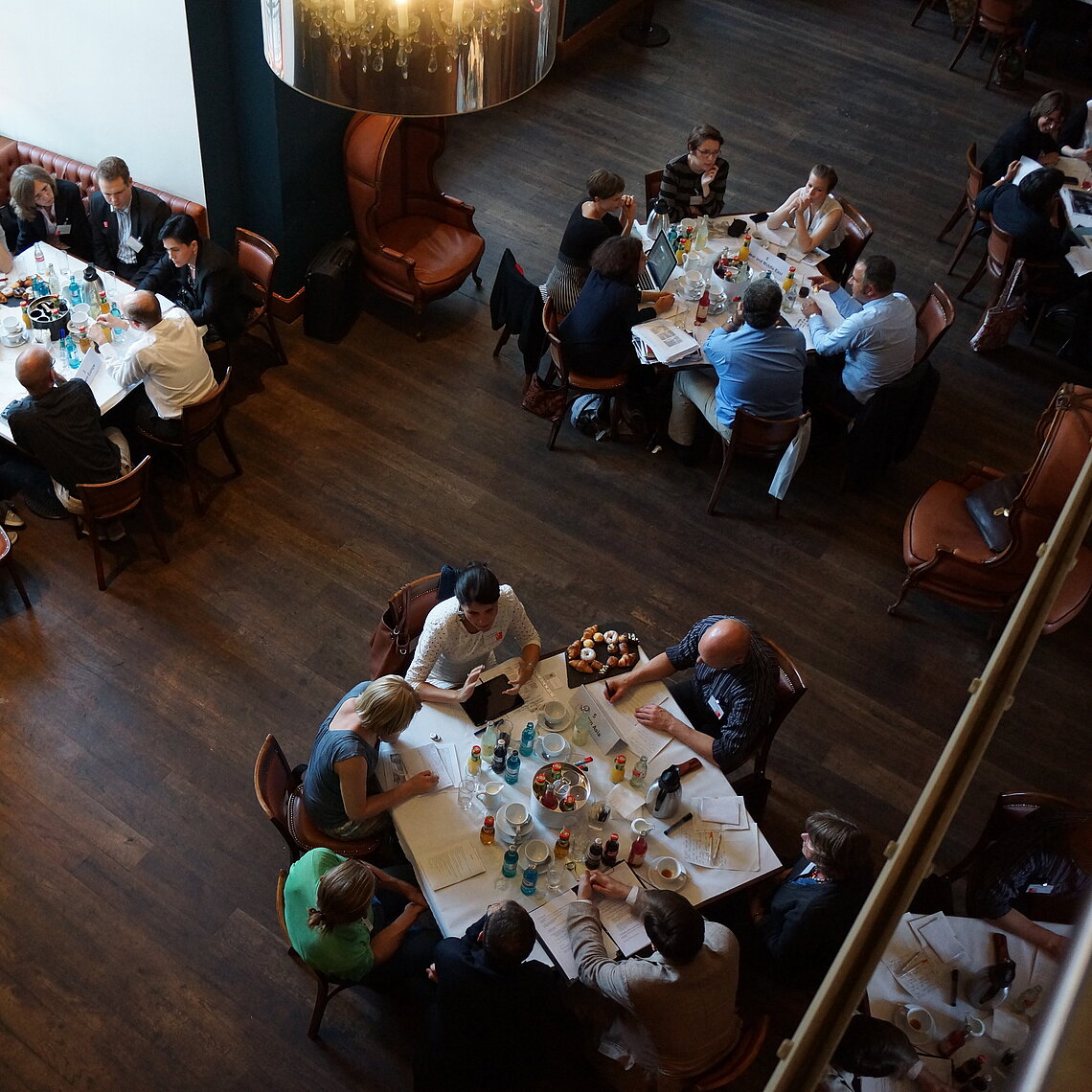 Mehrere Gruppen von Menschen sitzen an Tischen in einem Raum mit Holzboden und großen Lampen, sie sind mit Getränken und Papieren beschäftigt.
