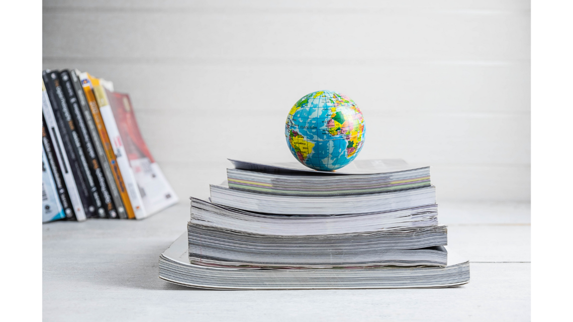 A small colorful globe sits on top of a stack of books or magazines on a white surface, with blurred books leaning on a shelf in the background.