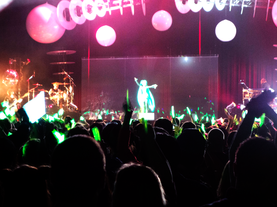 Konzertszene mit einer großen Menschenmenge im Vordergrund, die grüne Leuchtstäbe schwenkt. Im Zentrum der Bühne erscheint ein holografischer Auftritt der virtuellen Popfigur Hatsune Miku, umgeben von farbig beleuchteten Kugeln und Musikern. Die Bühne ist in kräftigen Farben und Lichtern inszeniert, was eine energetische Atmosphäre erzeugt.