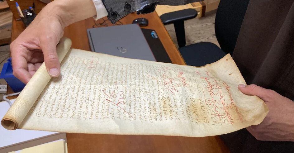 Person hält eine alte Schriftrolle mit handgeschriebenem Text und roten Markierungen über einem Holztisch.