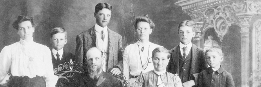 Schwarz-weiß Foto von acht Personen in historischer Kleidung. die vor einer dekorativen Wand mit Säulendetail für ein Gruppenporträt posieren.